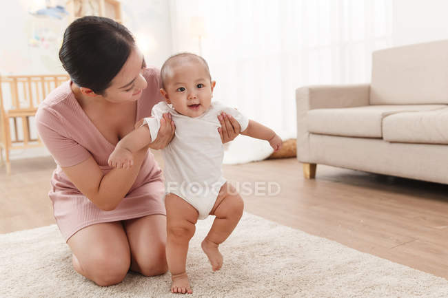 Молодая мать носит очаровательного младенца, ходящего по ковру — стоковое фото