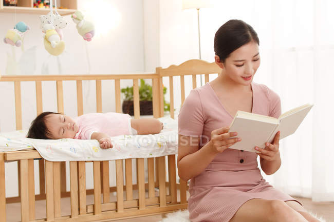 Young asian mother reading book while cute baby sleeping in crib — Stock Photo