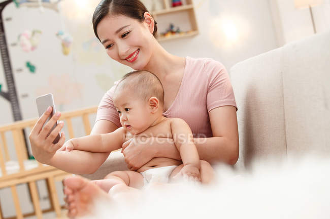 Sonriente madre joven abrazando al bebé y utilizando el teléfono inteligente en casa - foto de stock
