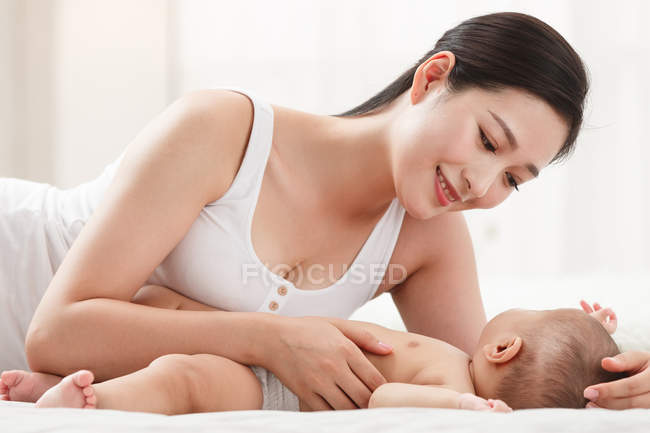 Усміхнена молода мати дивиться на чарівну дитину, що лежить на ліжку — стокове фото
