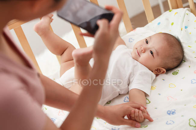 Recortado disparo de la madre joven sosteniendo teléfono inteligente y fotografiando bebé adorable acostado en la cuna - foto de stock