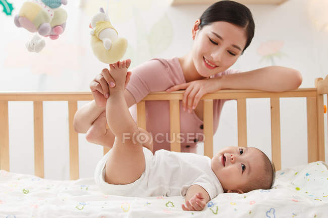 Smiling young mother playing with adorable infant baby lying in crib — Stock Photo