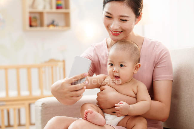 Счастливая молодая мама обнимает ребенка и делает селфи со смартфоном дома — стоковое фото