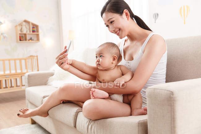 Усміхнена молода мати обіймає дитину і використовує смартфон вдома — стокове фото