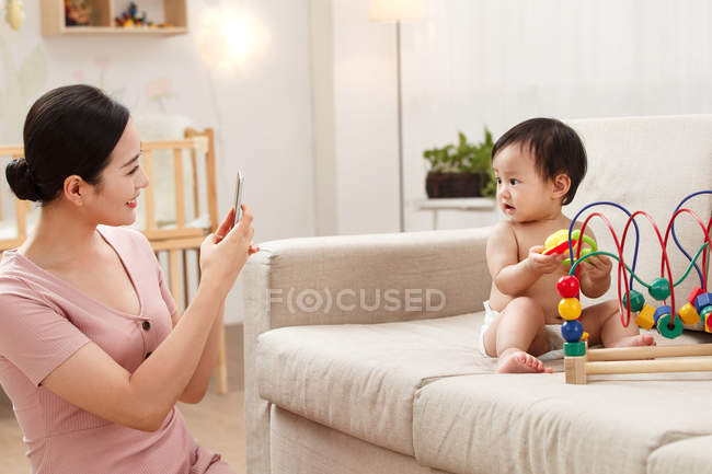 Sonriente joven madre sosteniendo teléfono inteligente y fotografiando adorable bebé jugando en el sofá - foto de stock