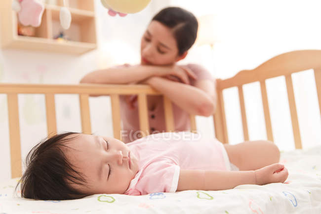 Madre joven cansada apoyada en una cuna de madera con los ojos cerrados mientras el bebé bebé duerme en primer plano, enfoque selectivo - foto de stock