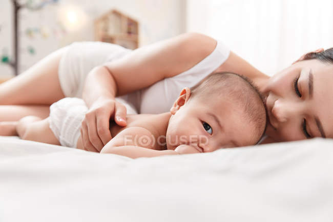 Молода мати лежить на ліжку з чарівною дитиною, дивлячись на камеру — стокове фото