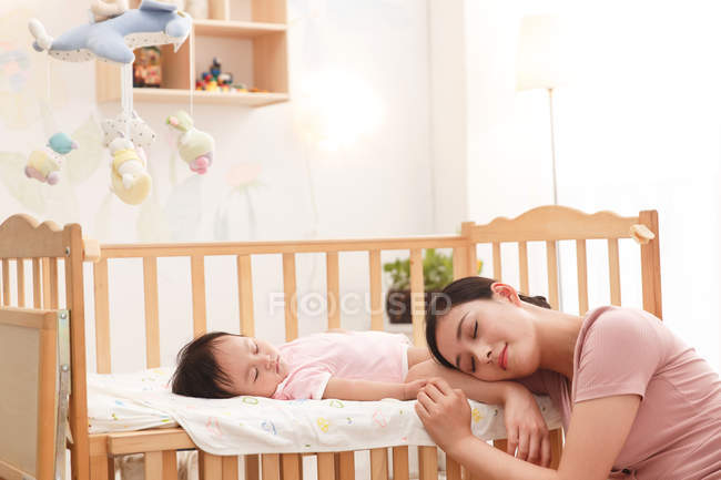 Fatigué jeune mère avec les yeux fermés penché à la crèche tandis que bébé adorable dormir — Photo de stock