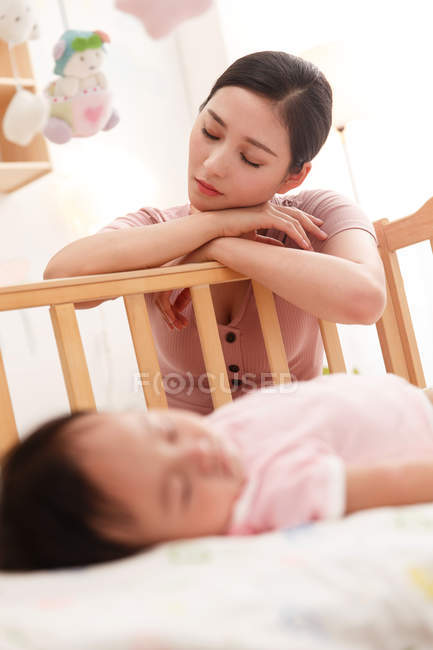 Müde junge Mutter lehnt mit geschlossenen Augen an Holzkrippe, während Säugling im Vordergrund schläft, selektiver Fokus — Stockfoto
