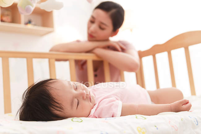 Close-up view of adorable infant child sleeping in crib, tired mother sitting behind — Stock Photo