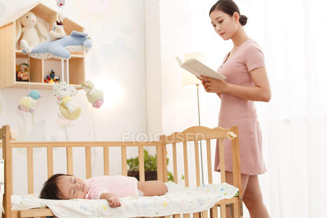 Young mother reading book while adorable infant child sleeping in crib — Stock Photo