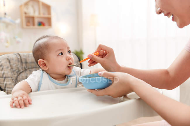 Plan recadré de souriant jeune mère nourrissant bébé adorable à la maison — Photo de stock