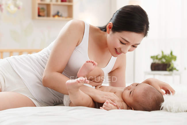 Lächelnde junge Mutter spielt mit entzückendem Baby, das auf dem Bett liegt — Stockfoto