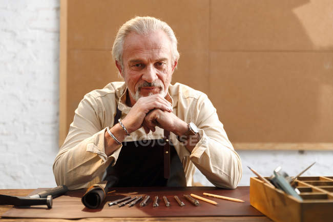 Серьезный зрелый мужчина в фартуке сидит за столом с инструментами и смотрит в камеру на рабочем месте — стоковое фото