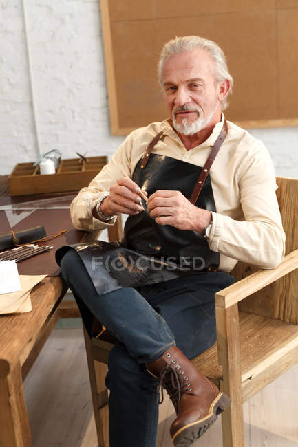 Ernster reifer Mann in Schürze sitzt mit Werkzeug am Tisch und blickt am Arbeitsplatz in die Kamera — Stockfoto