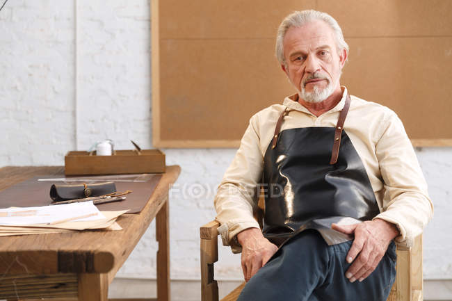 Serio uomo maturo in grembiule seduto a tavola con gli strumenti e guardando la fotocamera sul posto di lavoro — Foto stock