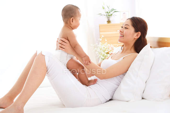 Vue latérale de la jeune mère asiatique jouant avec adorable petit bébé sur le lit — Photo de stock