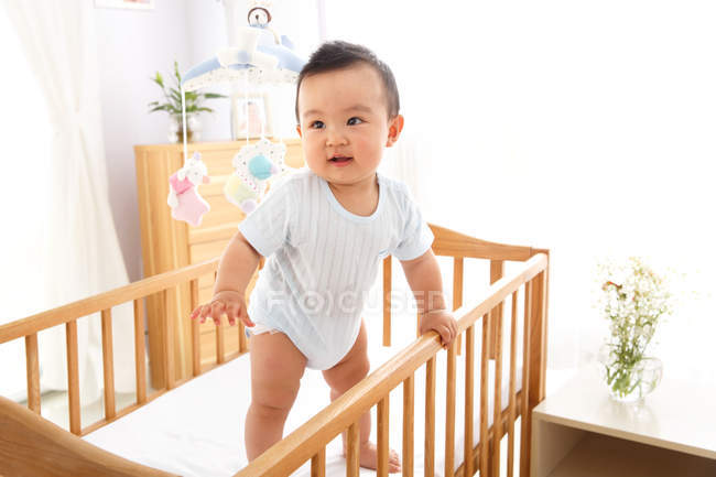 Милый азиатский мальчик стоит на кроватке и смотрит в сторону — стоковое фото