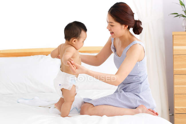 Vista lateral de jovem asiático mãe brincando com adorável pouco bebê na cama — Fotografia de Stock