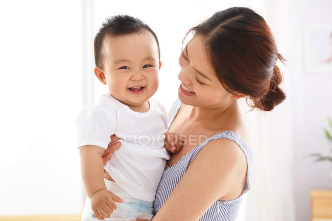Feliz jovem mãe segurando adorável bebê alegre em casa — Fotografia de Stock