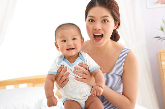 Glückliche junge Mutter mit entzückendem Baby, das zu Hause in die Kamera schaut — Stockfoto