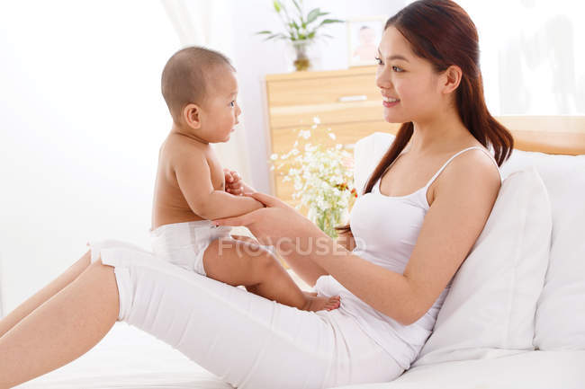 Вид сбоку на молодую мать, играющую с очаровательным маленьким ребенком на кровати — стоковое фото