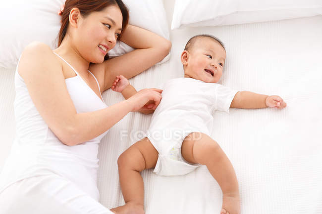 Blick aus der Vogelperspektive auf eine glückliche junge Mutter, die mit einem entzückenden Baby spielt, das auf dem Bett liegt — Stockfoto