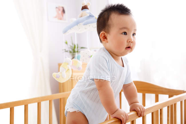 Очаровательный любопытный китайский ребенок стоит в кроватке и смотрит в сторону — стоковое фото