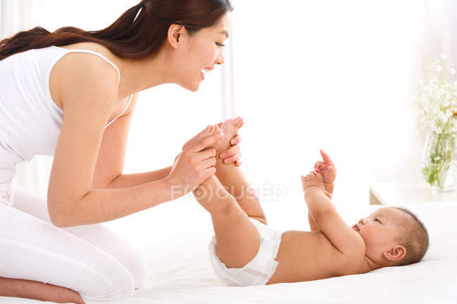 Happy young mother holding feet and playing with adorable little baby at home — Stock Photo
