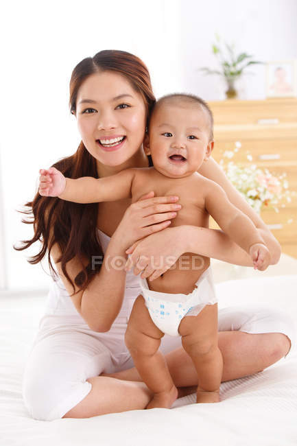 Feliz joven madre abrazando lindo bebé y sonriendo a la cámara - foto de stock