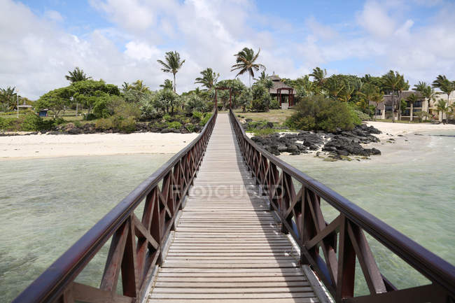 Деревянный мост через море и красивый песчаный пляж со зданиями и пальмами — стоковое фото