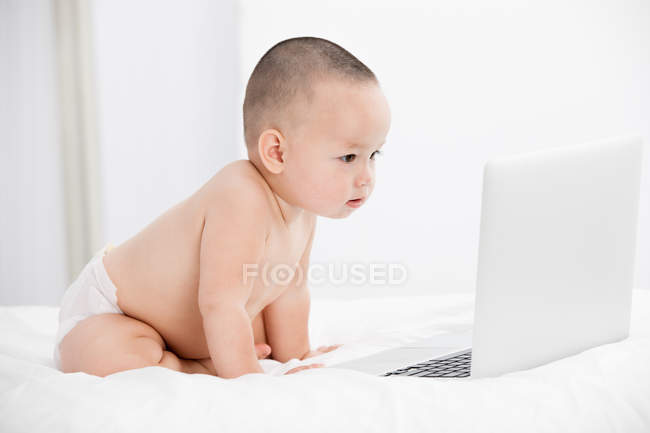 Bebê adorável em fralda sentado na cama e olhando para o computador portátil — Fotografia de Stock