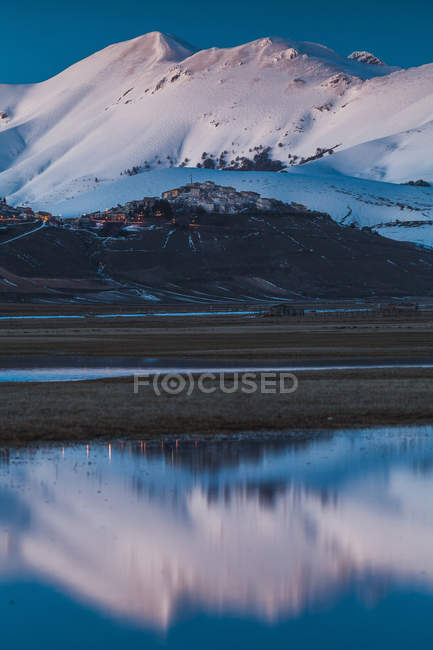 A view of Castelluccio di Norcia in front of a big snowed mountaind and reflected in a lake in the valley, Umbria, Italy — Stock Photo