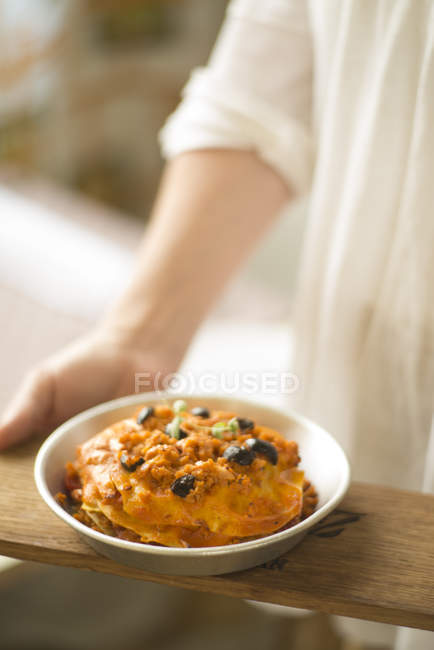 Female chef holding bowl of lasagna with soy sauce and olives. — Stock Photo