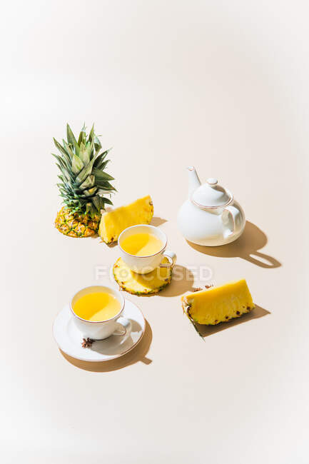 Tazas llenas de jugo de piña tropical fresco con fruta de piña en rodajas y tetera sobre fondo claro - foto de stock