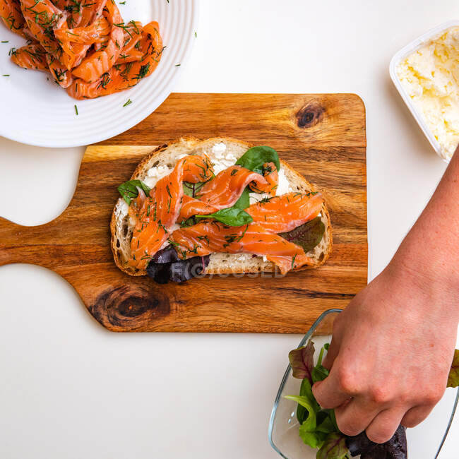 Vista de manos femeninas cocinando una bruschetta con salmón, tomate y verduras sobre fondo blanco - foto de stock