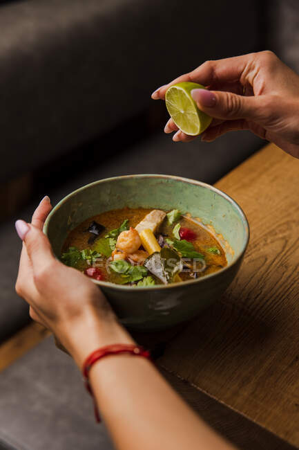 Frauenhände pressen Limetten zu Suppe mit Garnelen, Gemüse und Fischstücken auf den Tisch — Stockfoto