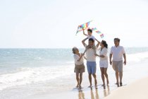Chico chino con cometa en los hombros del padre caminando con la familia en la playa - foto de stock