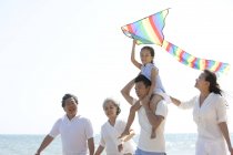 Китаянка с воздушным змеем на плечах отца гуляет с семьей на пляже — стоковое фото