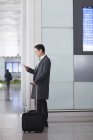 Homem asiático usando smartphone com bagagem de rodas no lobby do aeroporto — Fotografia de Stock