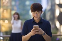 Young Chinese man using smartphone with woman on the phone in background — Stock Photo