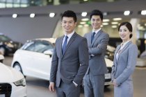 Confident salespeople standing with new cars in showroom — Stock Photo
