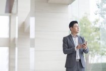 Азиатский бизнесмен стоит перед стеклянной дверью со смартфоном — стоковое фото