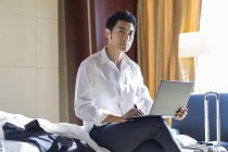 Hombre de negocios chino usando el ordenador portátil en la cama en la habitación de hotel - foto de stock