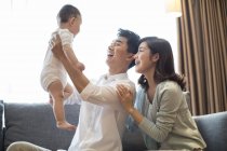 Padres chinos sosteniendo al niño y sonriendo en el sofá - foto de stock