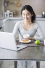 Mulher asiática usando laptop enquanto toma café da manhã — Fotografia de Stock
