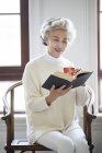 Senior Chinese woman reading book with cup of tea — Stock Photo