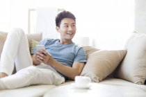 Homem chinês segurando smartphone e olhando para longe no sofá — Fotografia de Stock