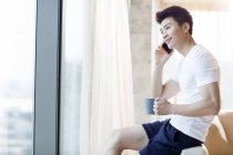 Chinese man with coffee talking on phone at home — Stock Photo