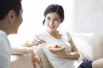 Chinese woman eating fruit salad and looking at man on sofa — Stock Photo